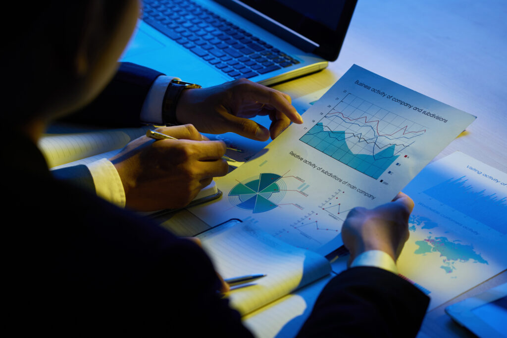 Duas pessoas de terno analisando relatórios com gráficos de atividade empresarial ao lado de um notebook