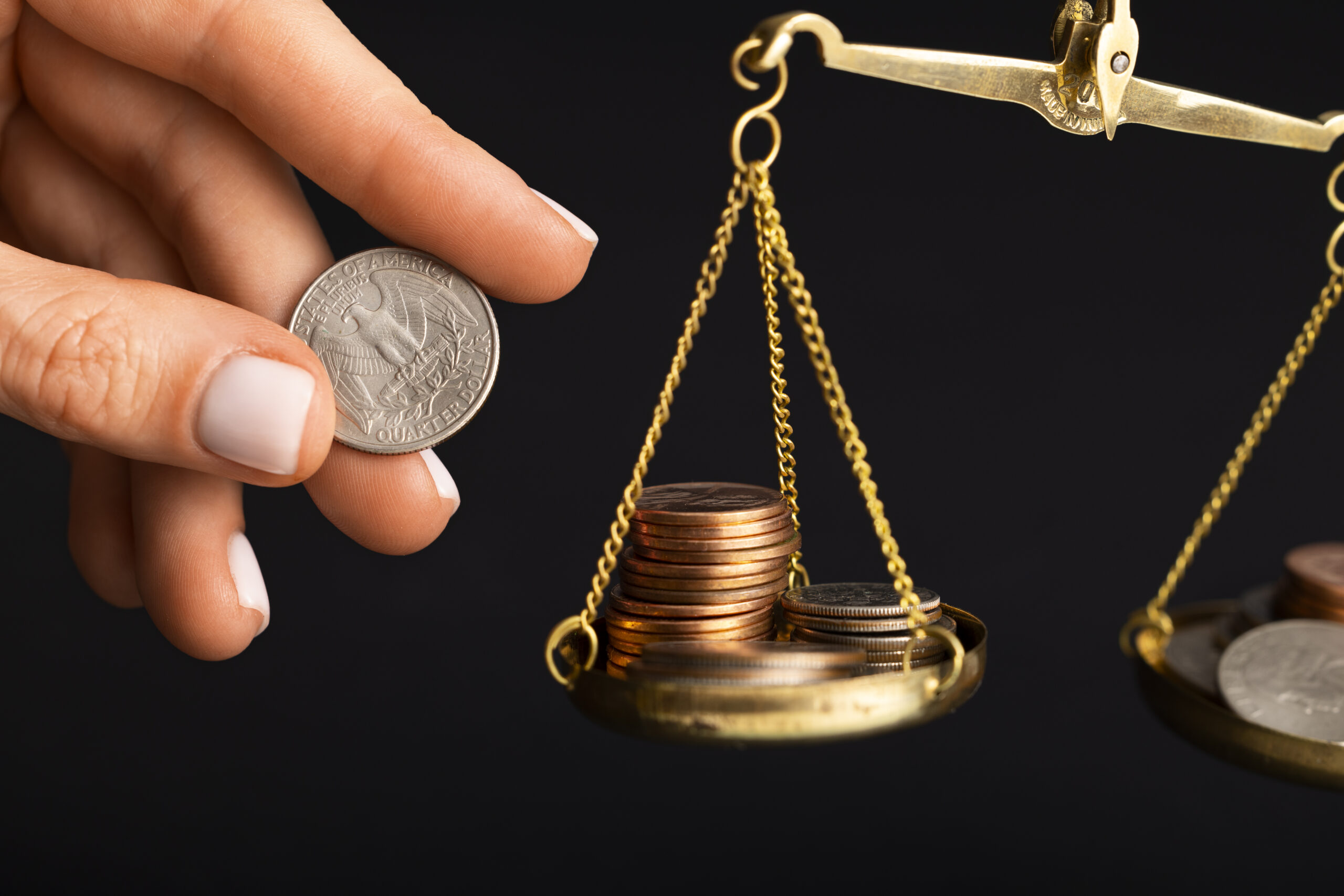 Mão segurando uma moeda de 25 centavos de dólar ao lado de uma balança dourada de precisão com pilhas de moedas